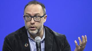 Jimmy Wales gestures with a microphone in his hand, set against a rich blue background