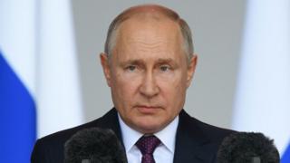 putin looking unhappy in front of a microphone with a pair of russian flags behind him