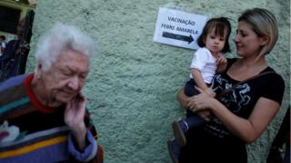 Idosa, mãe e bebê são fotografadas perto de placa indicando local de vacinação