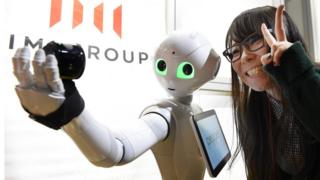A woman poses as humanoid robot Pepper takes a picture at a booth of digital marketing company IMJ Corporation during the Pepper App Challenge 2015 in Tokyo on February 22, 2015. Mobile phone carrier Softbank, which developed the humanoid robot, held the application competition for Pepper on February 22.