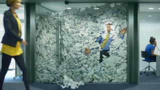A businessman is trapped in his glass office by a surplus of discarded ideas on paper . His female colleague in the next office is working more efficiently and is oblivious to him being trapped, as is a passing female office worker.