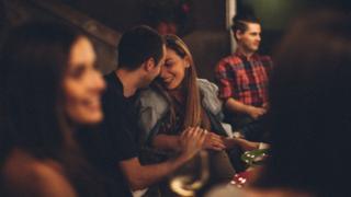 Una pareja coquetea en un ambiente social. Él tiene la mano sobre su mano y ella se está riendo.
