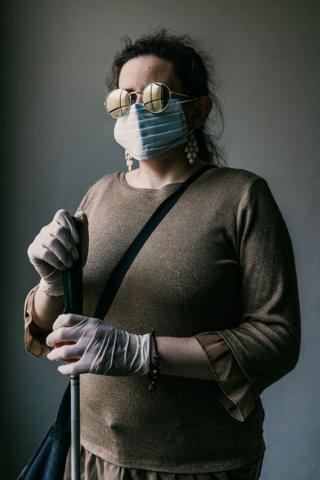 A blind woman stands with her walking cane whilst wearing a face mask and gloves