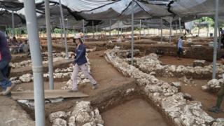 Plastic coverings hang over the excavation site as workers walk along stone-lined paths