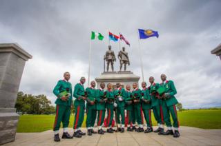 Male and female soldiers in green and red uniforms stand in front of the memorial featuring flags and two statues in Abuja