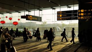 Passengers with bags and trolleys walking under signs for gates in Terminal 3 Heathrow