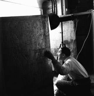 Howard Carter peering into the burial chamber from an opening in a wall engraved with hieroglyphs