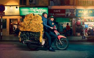 Two men pose on a motorcycle with a large mound of yellow flowers on the back
