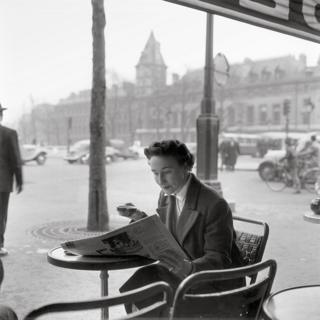 A woman drinks a coffee and reads a newspaper as she sits outside a cafe