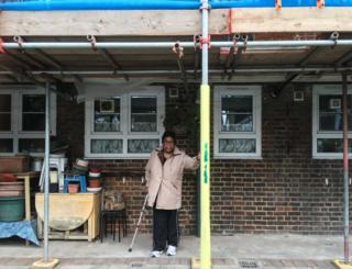 A woman with a stick stands next to a residential building, looking to the camera