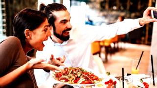 Happy couple at a restaurant, taking a "selfie" of themselves holding out a plate of food and smiling manically