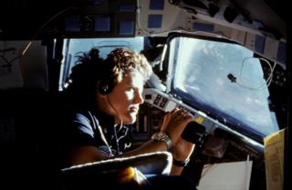 La astronauta Kathryn D. Sullivan, especialista en misiones 41G, usa binoculares para ver la Tierra a través de las ventanas de la cabina delantera.