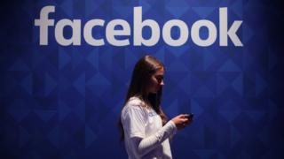 Una joven con un teléfono y con el cartel de Facebook de fondo.