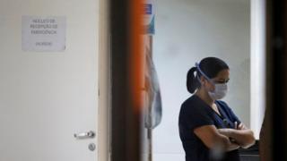 Enfermeira com braços cruzados e máscara olha para baixo dentro de sala de hospital