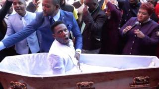Pastor Alph Lukau stands next to the coffin and places his hand on the stomach of the man he is claiming to resurrect.