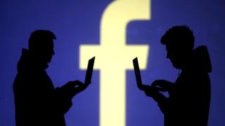 Sombra de duas pessoas mexendo em laptops à frente de grande logomarca do Facebook