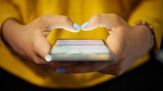 Close up of woman's hands, holding a smartphone - she's wearing a yellow top and is tapping on her phone's screen