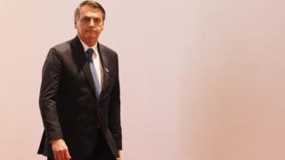 Bolsonaro aparece de terno e em pé na frente de fundo branco