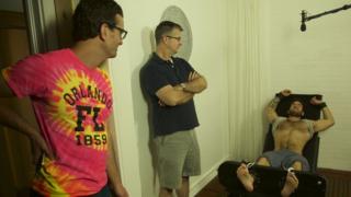 Un hombre sin camisa y descalzo está acostado en una silla rodeado de otros dos hombres que lo ven.