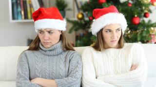 Two women wearing Santa hats looking away from each with a Christmas tree in the background