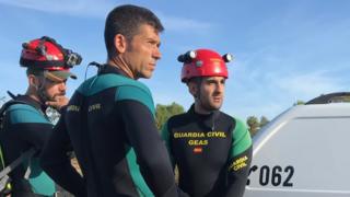 Three men in diving suit and helmet, sporting the logo of the Spanish Civil Guard, stand near their vehicle and look at the midfield behind the frame.