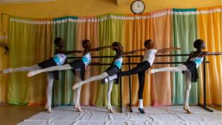 Young dancers dressed in matching leotards hold a ballet barre during a class.