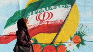 Mulher com véu passa em frente a pintura na parede da bandeira do Irã