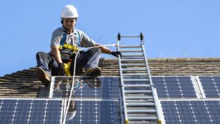 A maintenance person uses a ladder and harnesses to install equipment around a solar panel array. UK