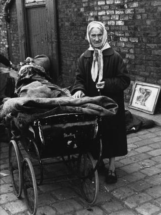 An elderly woman stands next to a pram with bundles of cloth on