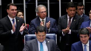 O senador Flávio Bolsonaro e o ministro Paulo Guedes (na foto, à esquerda e em pé) e o presidente da Casa, Davi Alcolumbre (sentado, ao centro), entre outros, comemoram aprovação de texto-base no Senado
