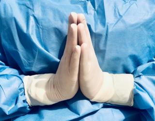 An NHS staff member with gown and gloves holds their hands in prayer