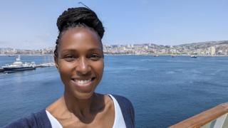 Brandee Lake smiling at the camera in a selfie, her braids are worn in a high bun and she is stood by a sea port.