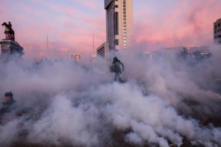 Police officer in riot gear runs amidst smoke bombs towards protesters