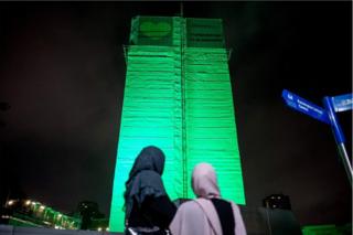 Grenfell Tower is lit with green light as people look on