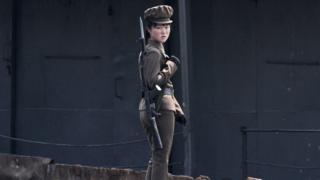 Северокорейская женщина-офицер в зеленой форме и пистолете смотрит на камеру