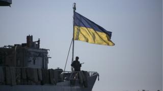 A Ukrainian navy ship, with a soldier standing in front of a Ukrainian flag