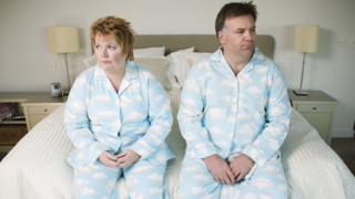 Hombre y mujer vestidos iguales y sentados en el borde de la cama con cara de descontento.