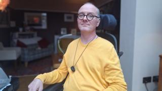 Glyn Jones sat in wheelchair in house wearing yellow sweater.