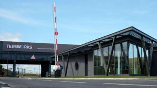 A red-and-white striped barrier outside the Teesworks entrance, a large grey building.