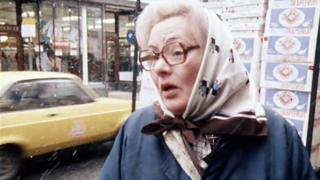 A older woman wearing a blue coat, patterned headscarf, and brown glasses speaking to the interviewer (out of sight)