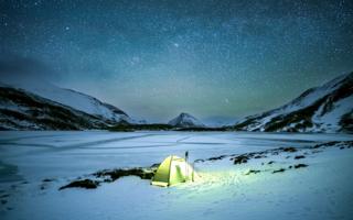 Tent brightly lit from inside sat on the snow below a star covered sky at night