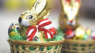 A chocolate Easter rabbit, wrapped in golden foil sits in a bed of green tissue paper, surrounded by little chocolate eggs.