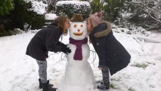 Two girls kissing a snowman on the cheek.
