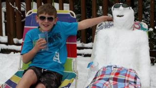 A boy on a sun lounger in sunglasses and shorts next to a snowman on a sun lounger.