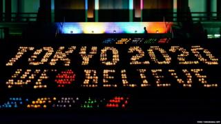 A man walks past an illuminated sign reading Tokyo 2020 We Believe which is made from 2,020 candles at Zojoji temple in Tokyo