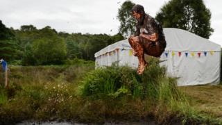 A man is jumping in the air , covered in mud, into the bog Jacuzzi.