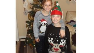 A boy and girl wearing Christmas jumpers by their tree