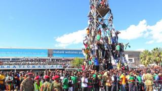 Thousands of Zambian people gather at Lusaka airport, some climbing on to a scaffold to get a glimpse of the football players