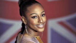 Alexandra Burke stands in front of a union flag. Her hair is tied up in a bun and she wears long, glamorous dangling earrings.