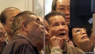 Пожилые инвесторы толпятся вокруг монитора цен на акции в брокерской компании в Гонконге
